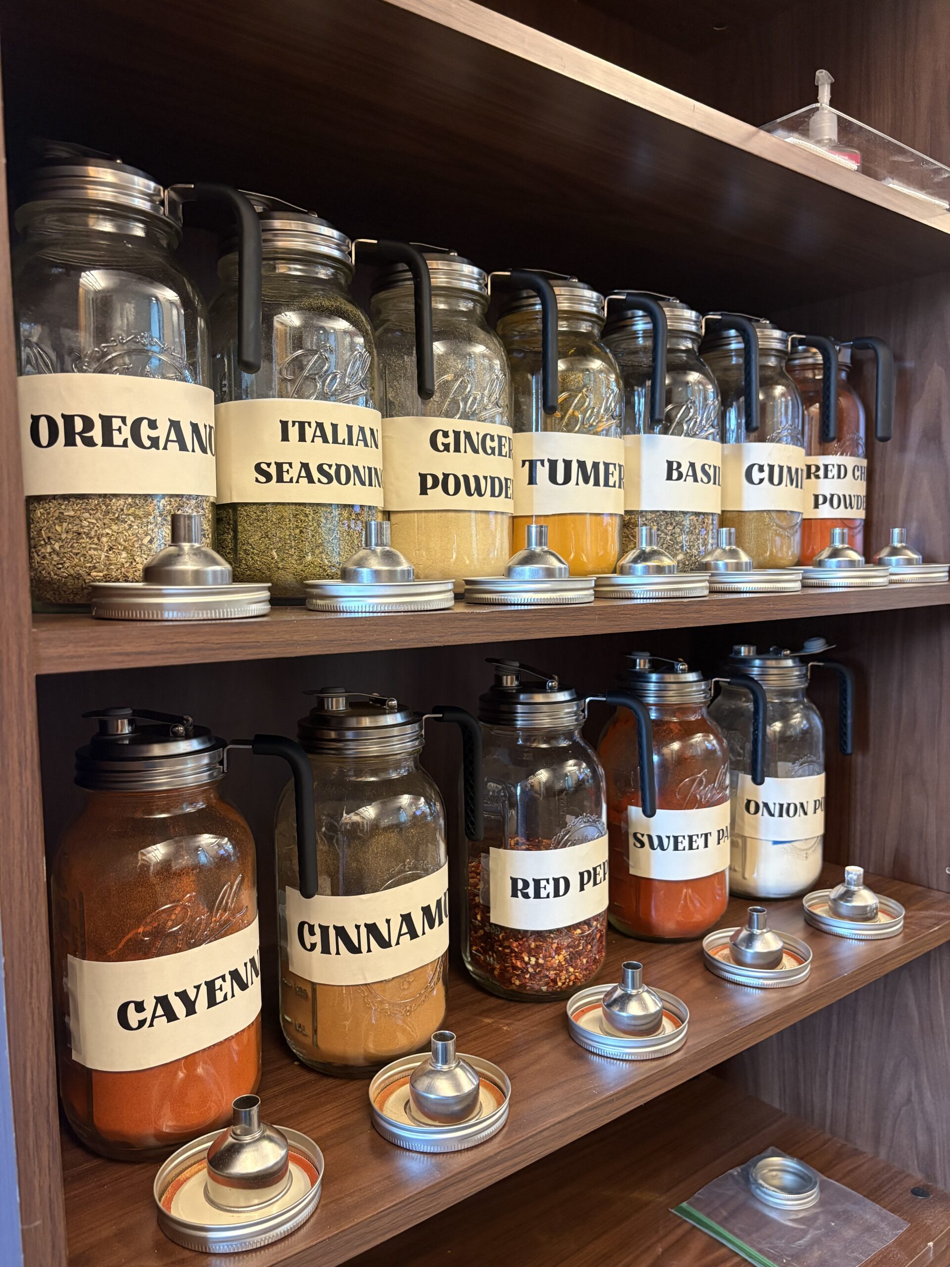 Two wooden shelves neatly lined with large glass Ball mason jars full of spices. Each jar is fitted with a black pour-handle lid and a bold, printed white label wrapped around the center. The top shelf contains jars labeled Oregano, Italian Seasoning, Ginger Powder, Turmeric, Basil, Cumin, and Red Chili Powder. The bottom shelf holds jars labeled Cayenne, Cinnamon, Red Pepper, Sweet Paprika, and Onion Powder, with small silver funnels and mason jar lids placed on the shelves in front of them.